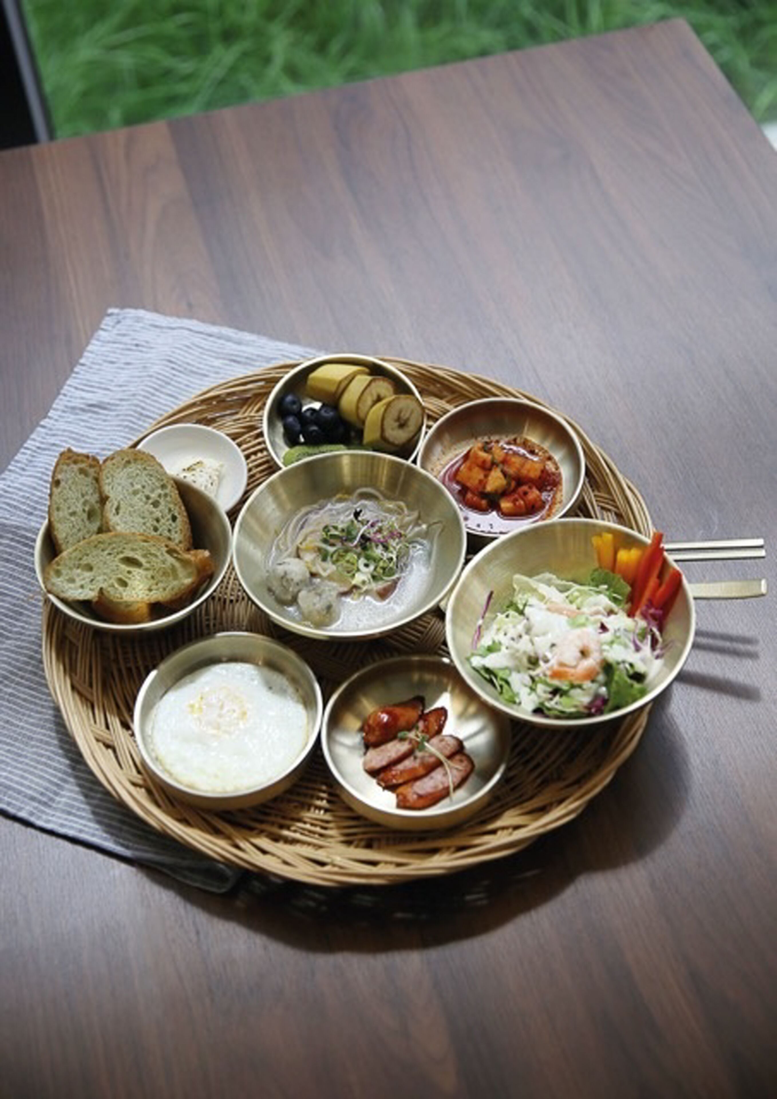 A wooden table with a woven tray holding various dishes including bread, salad, and soup.