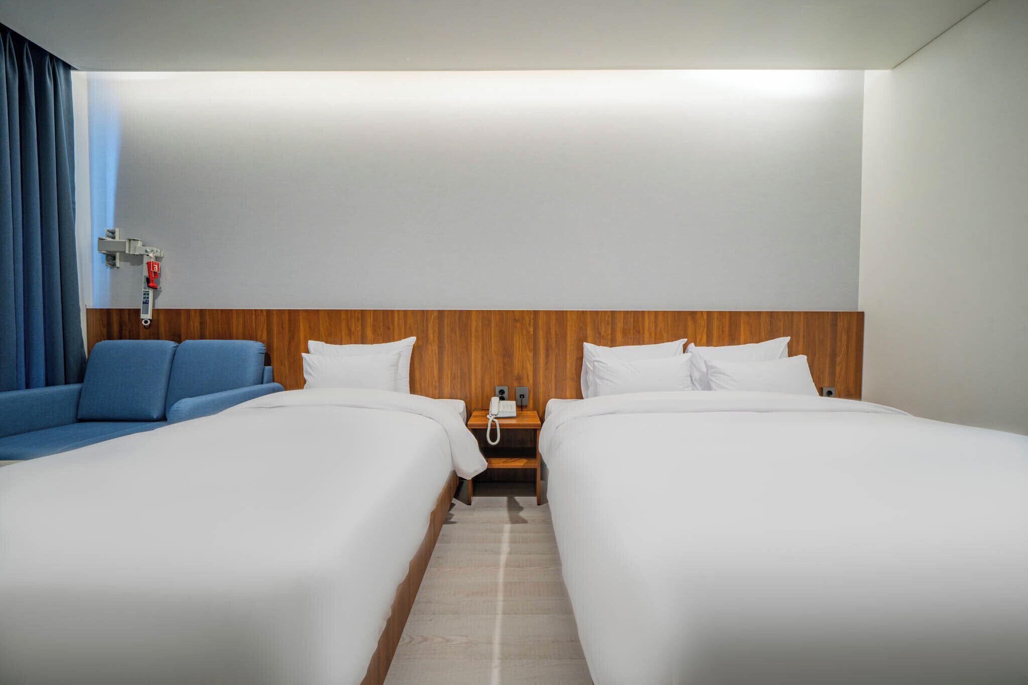 Una habitación de hotel con dos camas, cabecero de madera, mesita de noche con teléfono y sofá azul.