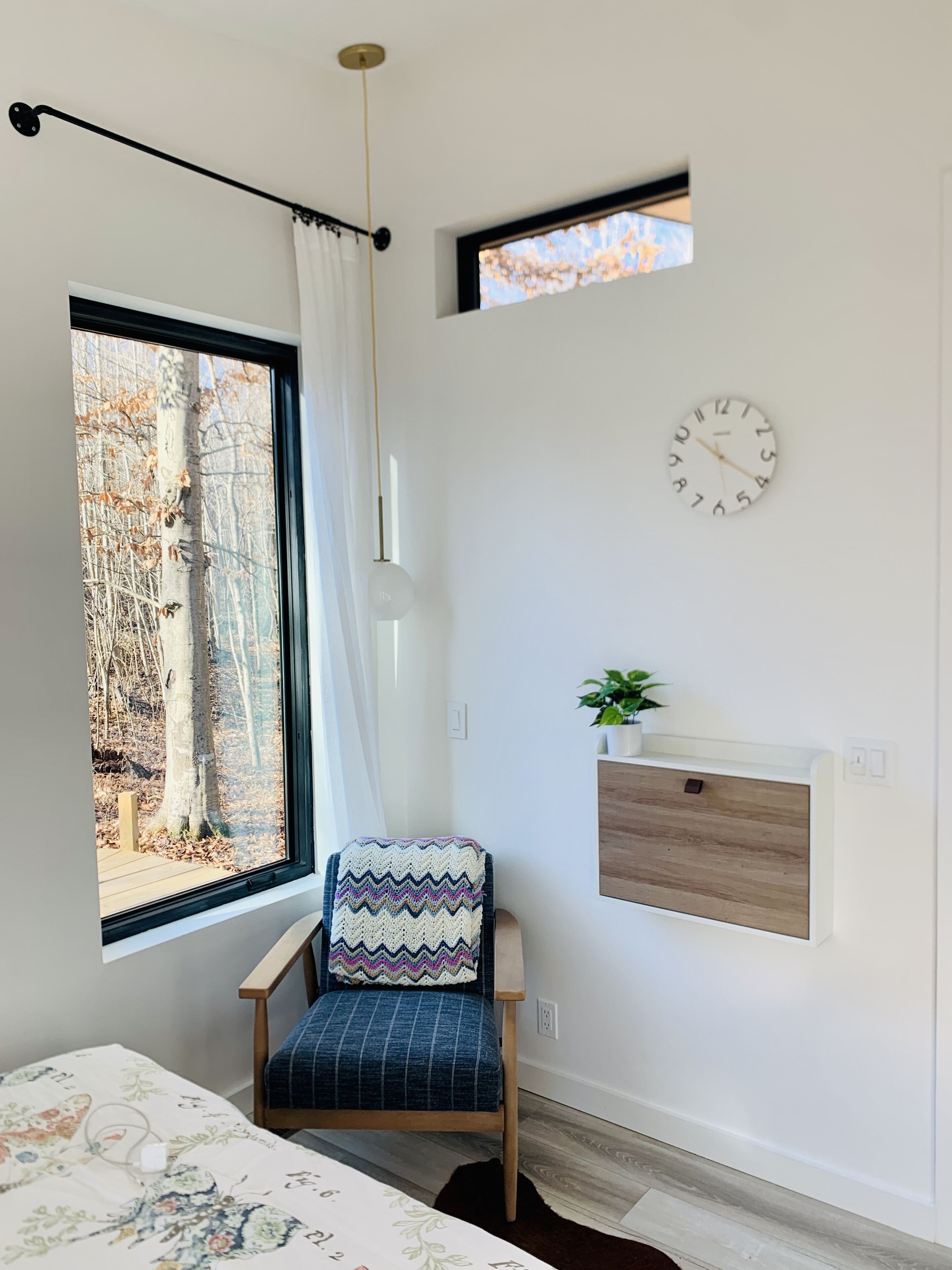 A modern bedroom with a large window, a wooden chair, a bedside table with a plant, and a wall clock.