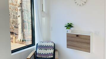 A modern bedroom with a large window, a wooden chair, a bedside table with a plant, and a wall clock.