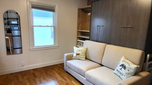 Una sala de estar con un sofá color beige, dos almohadones decorativos, piso de madera, una ventana, un armario empotrado de madera, y una estantería con microondas y cafetera.
