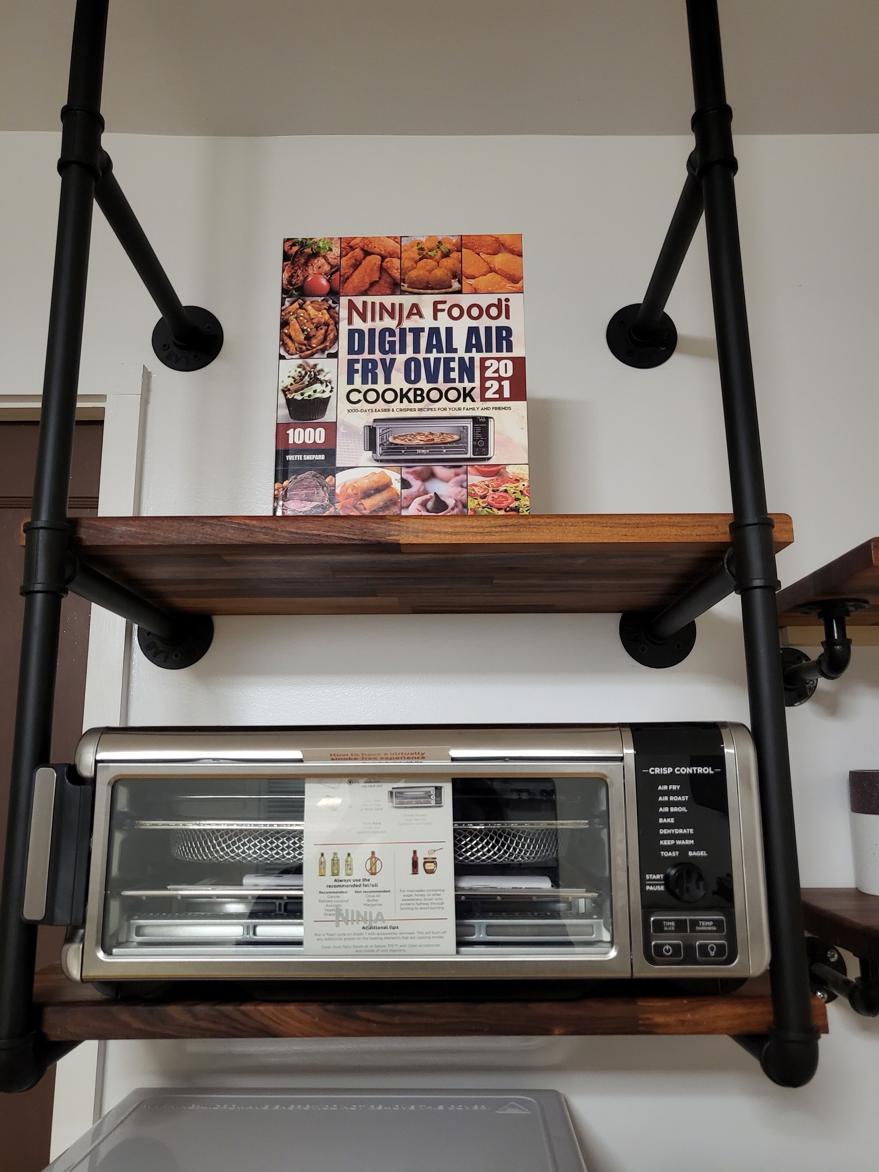 Une friteuse à air numérique est posée sur une étagère, et un livre de recettes intitulé « Ninja Foodi Digital Air Fry Oven 2021 » se trouve juste au-dessus.