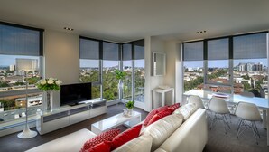 Una sala moderna con un televisor grande, sofás blancos, cojines rojos, una mesa de centro y vistas de un paisaje urbano a través de amplias ventanas.