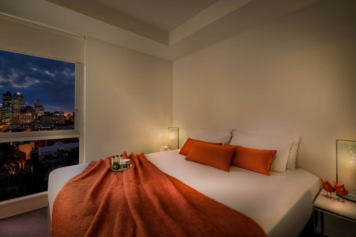 Habitación de hotel con una cama grande, ropa de cama naranja, una mesita de noche con lámpara, una ventana con vistas a la ciudad y una pequeña mesa con una vela y un adorno.