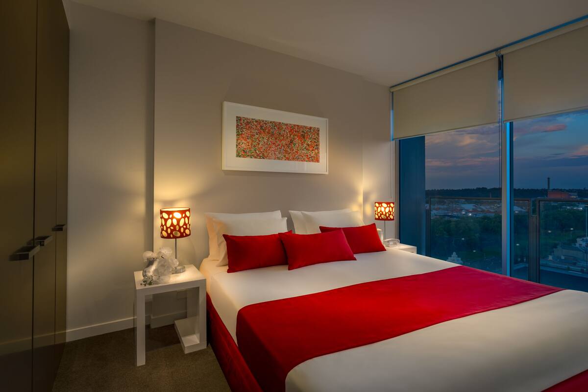 Una habitación de hotel moderna con una cama grande, ropa de cama roja, vista a la ciudad por la ventana y una obra de arte enmarcada en la pared.