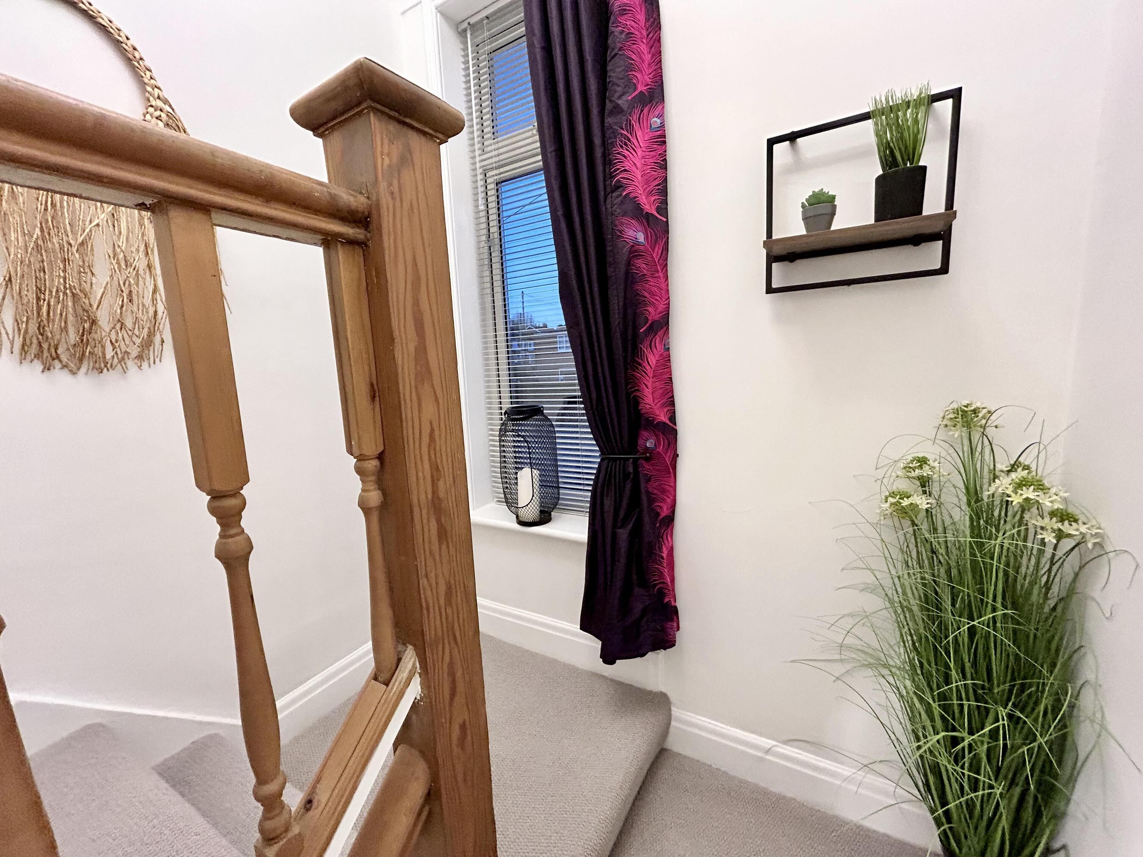 A staircase with wooden handrails, a window with blinds, a dark curtain, and a small shelf with plants.