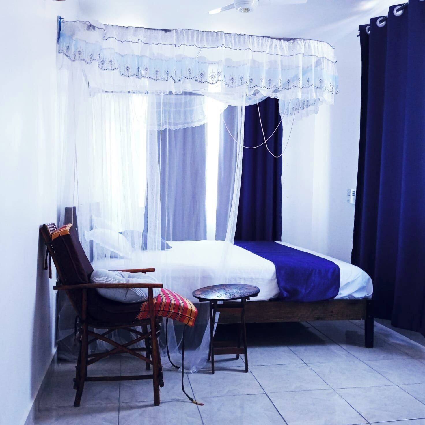 Un dormitorio con una cama, una silla, una mesita y una ventana con cortinas ligeras.