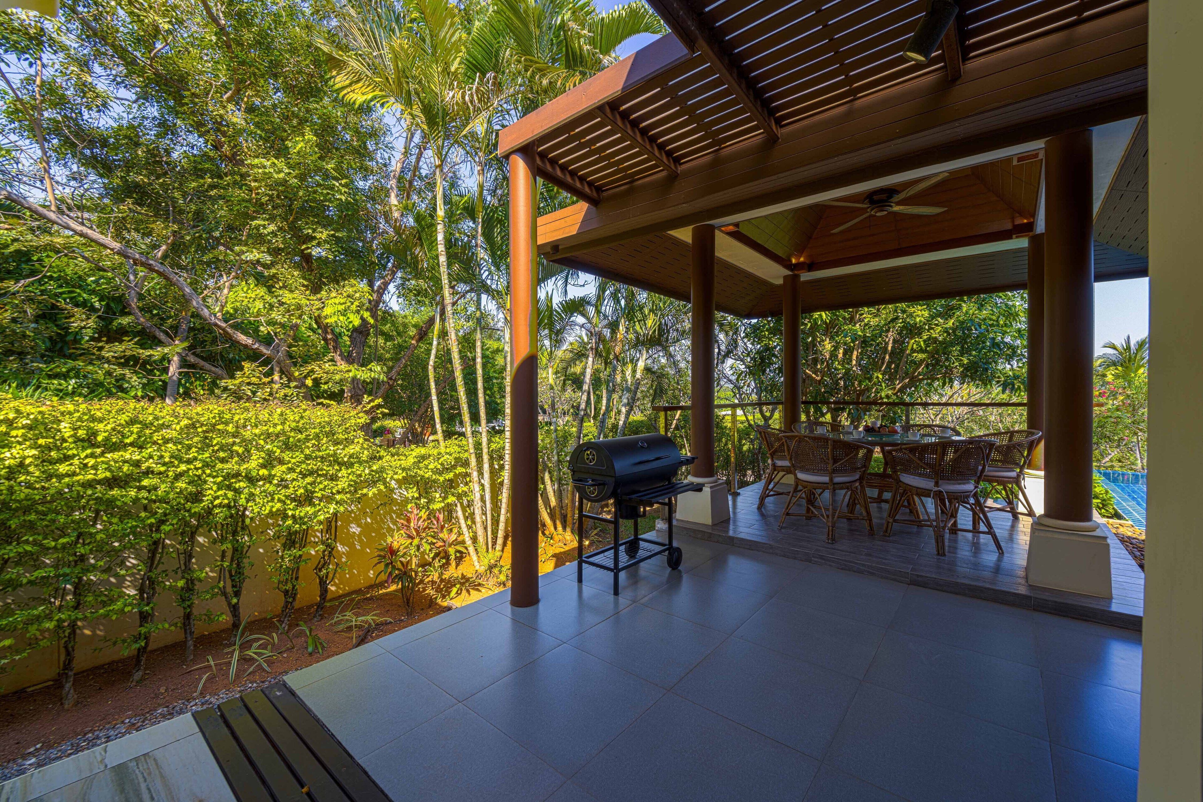 An outdoor patio with a grill, dining table, and chairs, surrounded by lush greenery and palm trees.