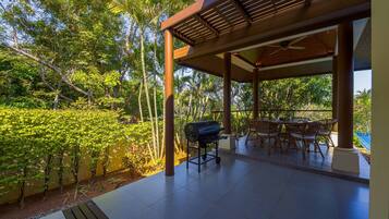 An outdoor patio with a grill, dining table, and chairs, surrounded by lush greenery and palm trees.