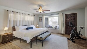 Ein Schlafzimmer mit einem Bett, Nachttischen, einem Deckenventilator, einem Fenster mit Vorhängen, einer Tür und einem stationären Ergometer.