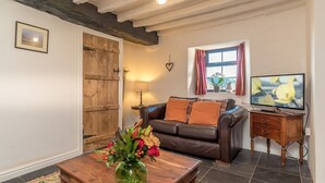 A living room with a brown leather sofa, a wooden coffee table with a vase of flowers, a television on a wooden stand, and a rustic wooden door.
