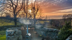 Eine rustikale, offene Sauna mit Steinsäulen und Holzkonstruktion, umgeben von einer Steinmauer und Bäumen, mit Sonnenuntergang im Hintergrund.