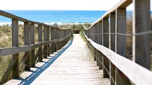 Ein hölzerner Steg führt zum Strand, begleitet von einer Metallreling auf einer Seite.