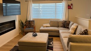 A living room with a sectional sofa, a flat-screen TV mounted on the wall, a fireplace, and a window with blinds.