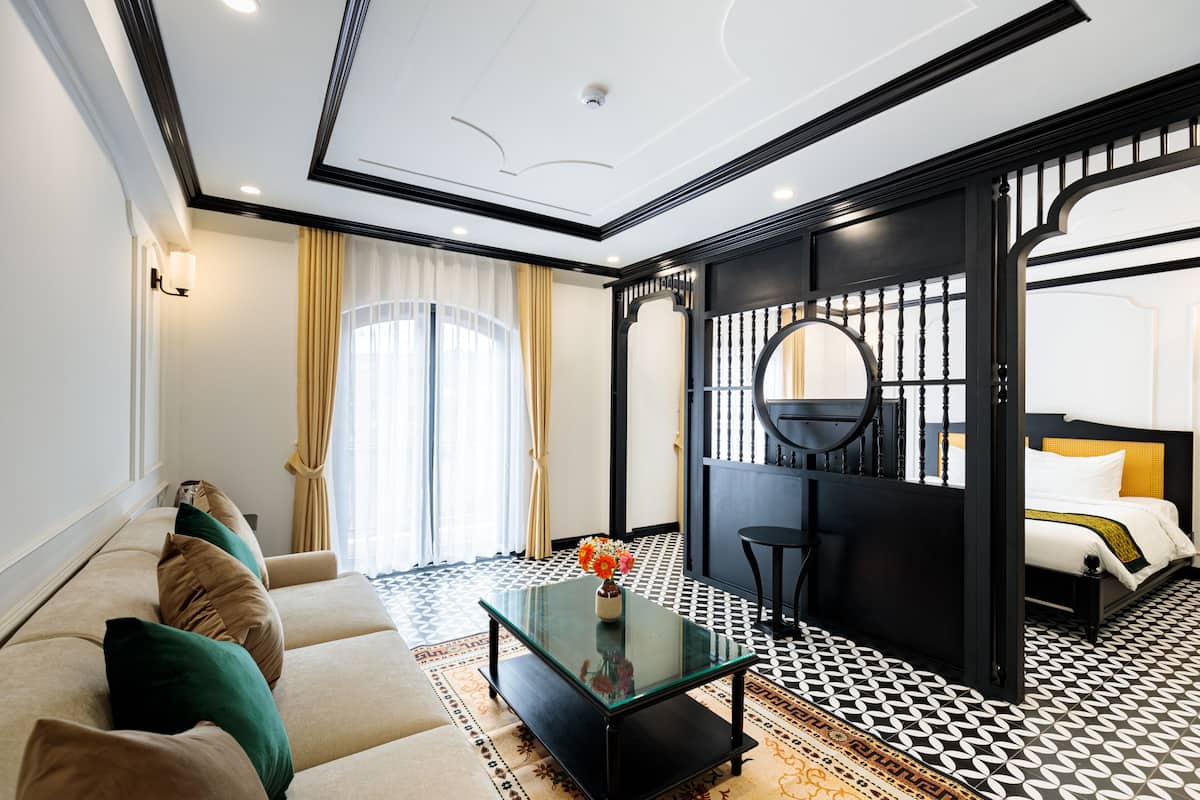 Ein Hotelzimmer mit einer Couch, einem Glastisch, einem Bett mit gelber Kopfteil und einer schwarzen Trennwand mit rundem Spiegel.