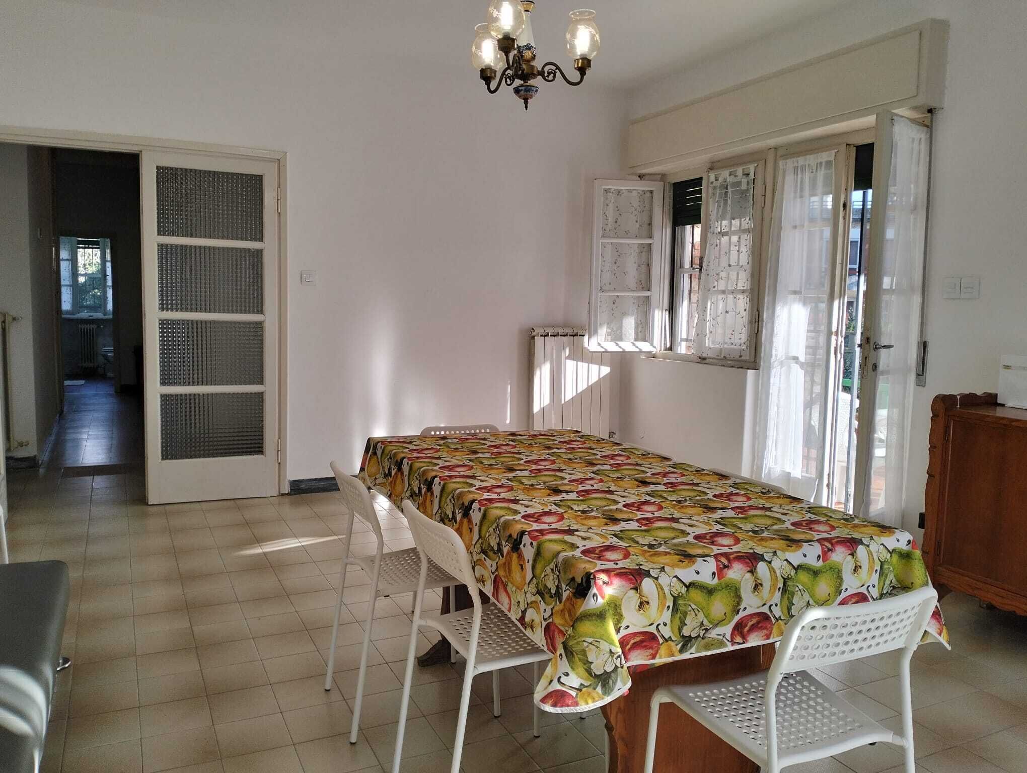 A dining area with a table covered by a fruit-patterned tablecloth, white chairs, and a chandelier.