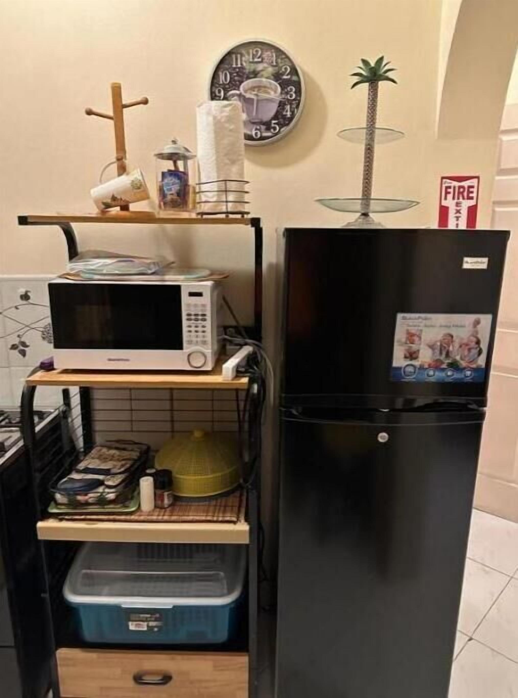 Une cuisine compacte équipée d’un four à micro-ondes, d’un réfrigérateur et d’une petite étagère sur laquelle se trouvent une horloge et un extincteur.