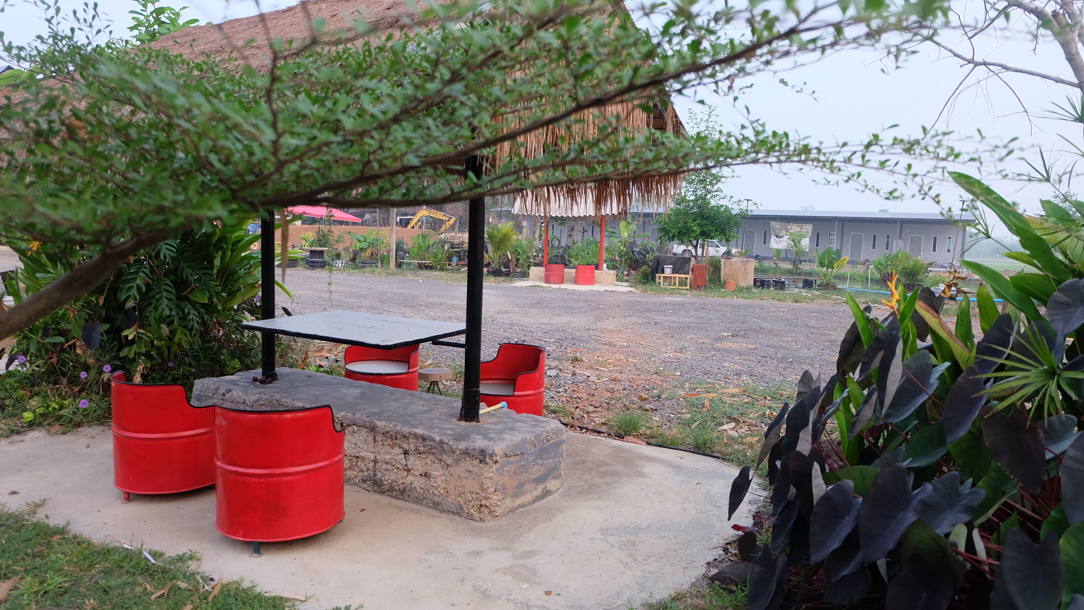 A covered outdoor seating area with a table and chairs, surrounded by greenery and a gravel path.