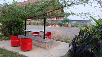 A covered outdoor seating area with a table and chairs, surrounded by greenery and a gravel path.