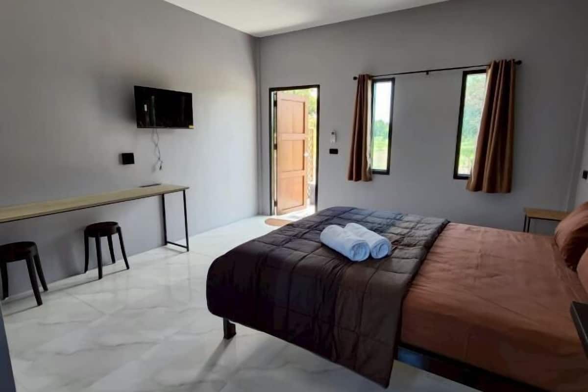Ein Schlafzimmer mit einem Bett, einem Schreibtisch, zwei Hocker, einem Fernseher und einem Fenster mit VorhÀngen.