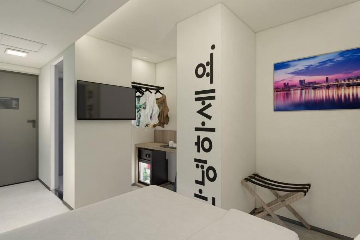 A hotel room with a bed, a TV mounted on the wall, a small desk with a computer, a coat rack, and a bench.