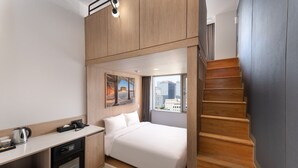 A compact hotel room with a bed, a small kitchenette, a desk, and a staircase.