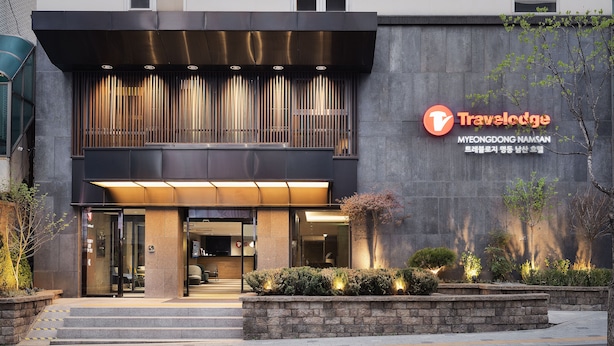 A modern building with a Travelodge sign, featuring a large entrance, steps leading up to the door, and a well-lit facade.