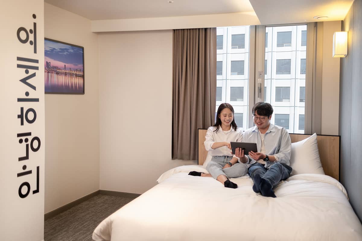 A hotel room with a bed, a window with curtains, a wall-mounted picture, and a bedside table.