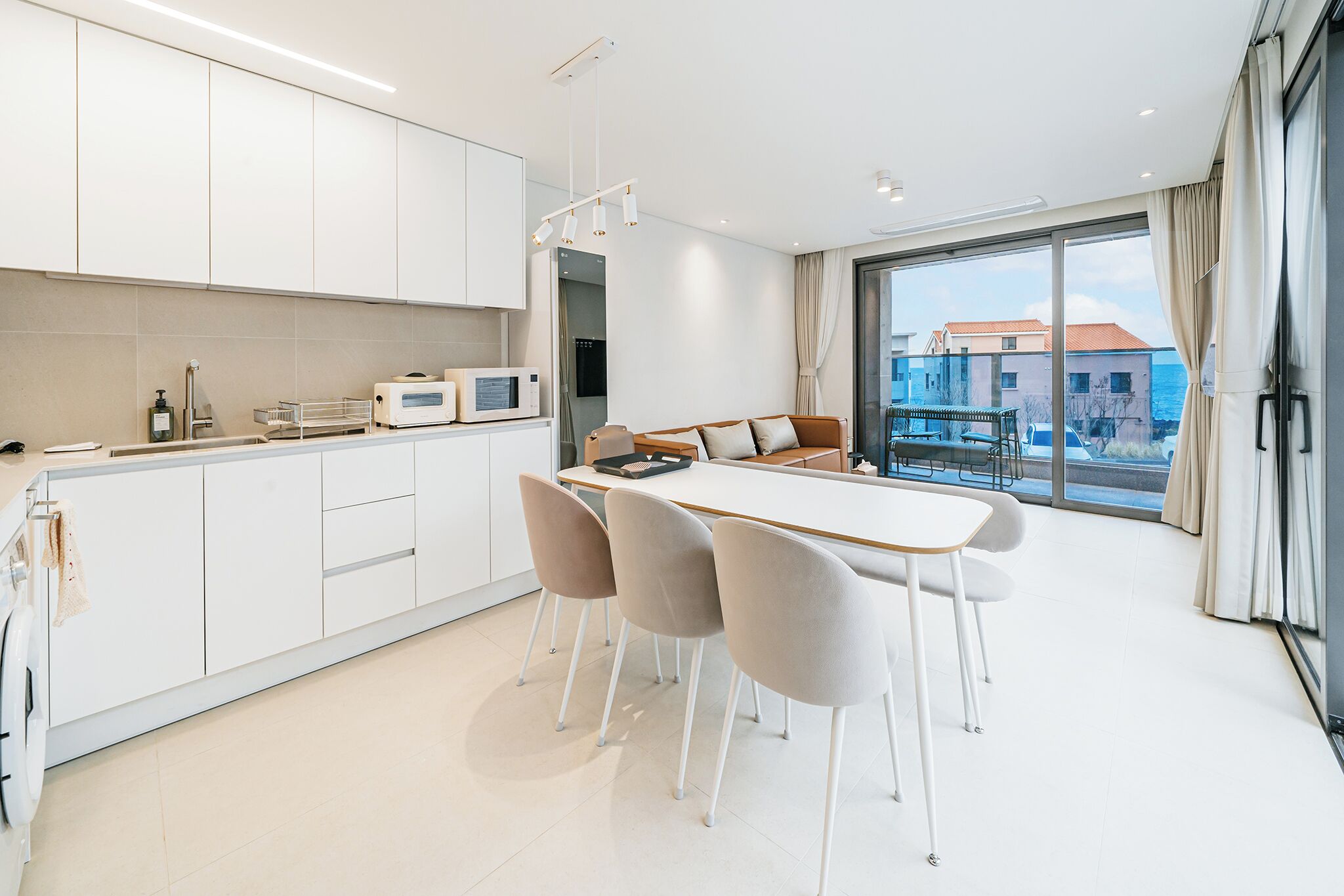 Une cuisine moderne avec des meubles blancs, un coin repas comprenant une table et des chaises, et une grande fenêtre offrant une vue sur les bâtiments et la mer.