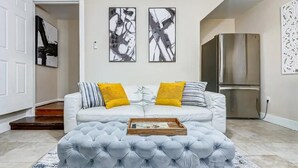 A living room with a white sofa, two yellow cushions, a grey tufted ottoman, and a wooden tray. There are abstract paintings on the wall and a stainless steel refrigerator.
