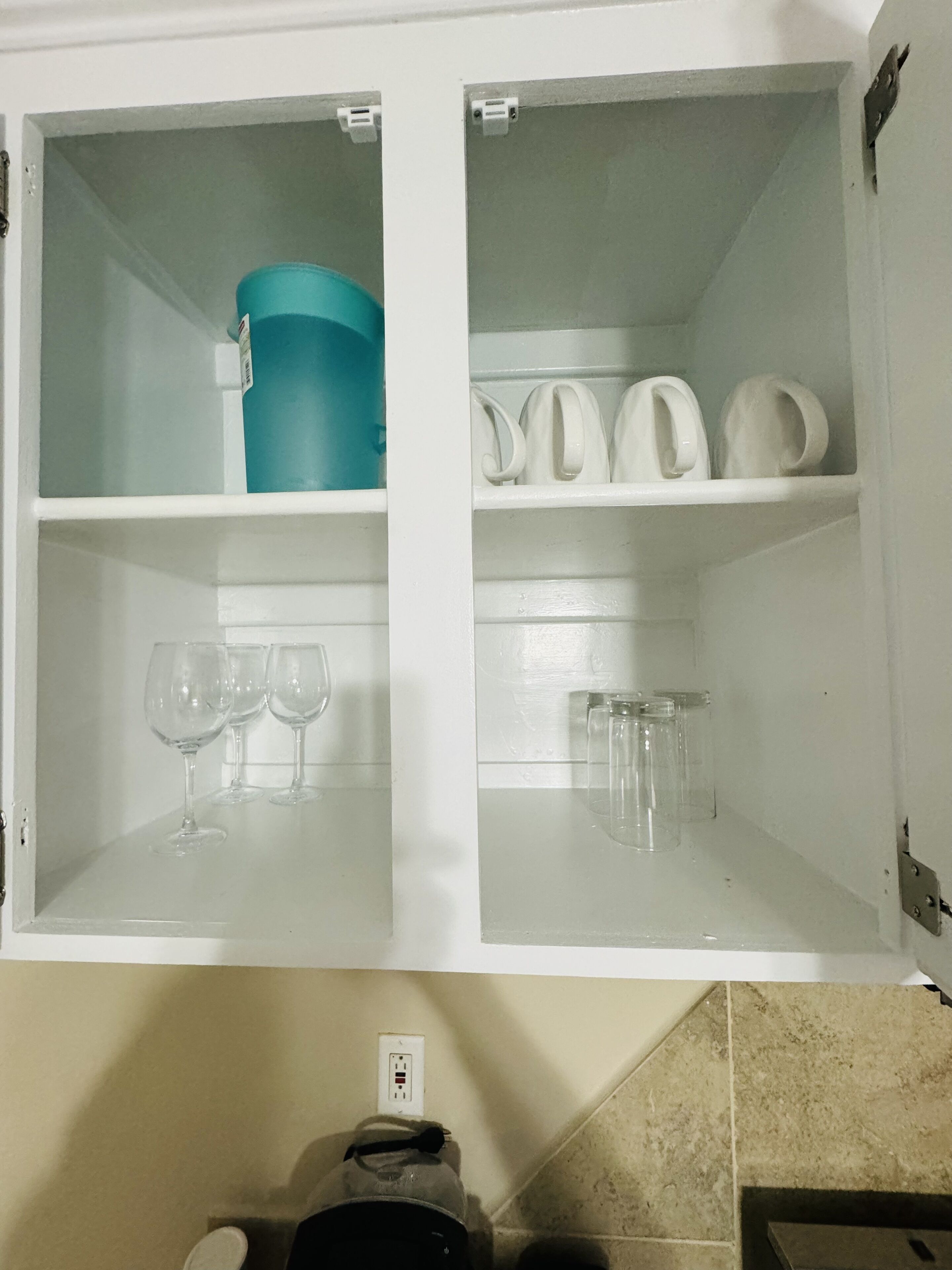 A white cabinet with two shelves; one shelf holds a blue container and wine glasses, and the other holds white dishes and glasses.