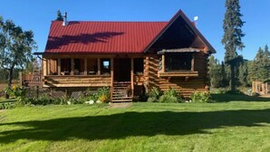 A log cabin with a red metal roof, a porch with outdoor seating, and a well-maintained lawn. - Log Cabin with Mountains and Lake View, enjoy relaxing in the hot tub (Clam Gulch)