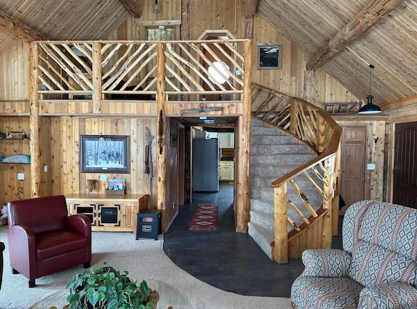 A rustic wooden interior with a high ceiling, a staircase, a red leather chair, a patterned sofa, and a television.