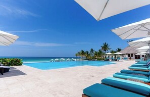 A poolside area with lounge chairs and umbrellas, a clear blue sky, and a view of the ocean.