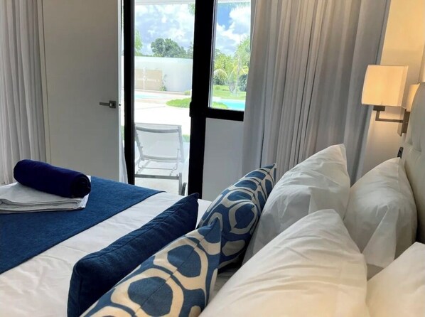 A hotel room with a bed, a window with curtains, a view of the outdoors, and a lamp on the wall.
