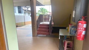A hallway with a staircase leading to an upper floor, a fire extinguisher on the wall, and a small table with a stool. - OYO 90915 Ilham Villa (Kota Bharu)