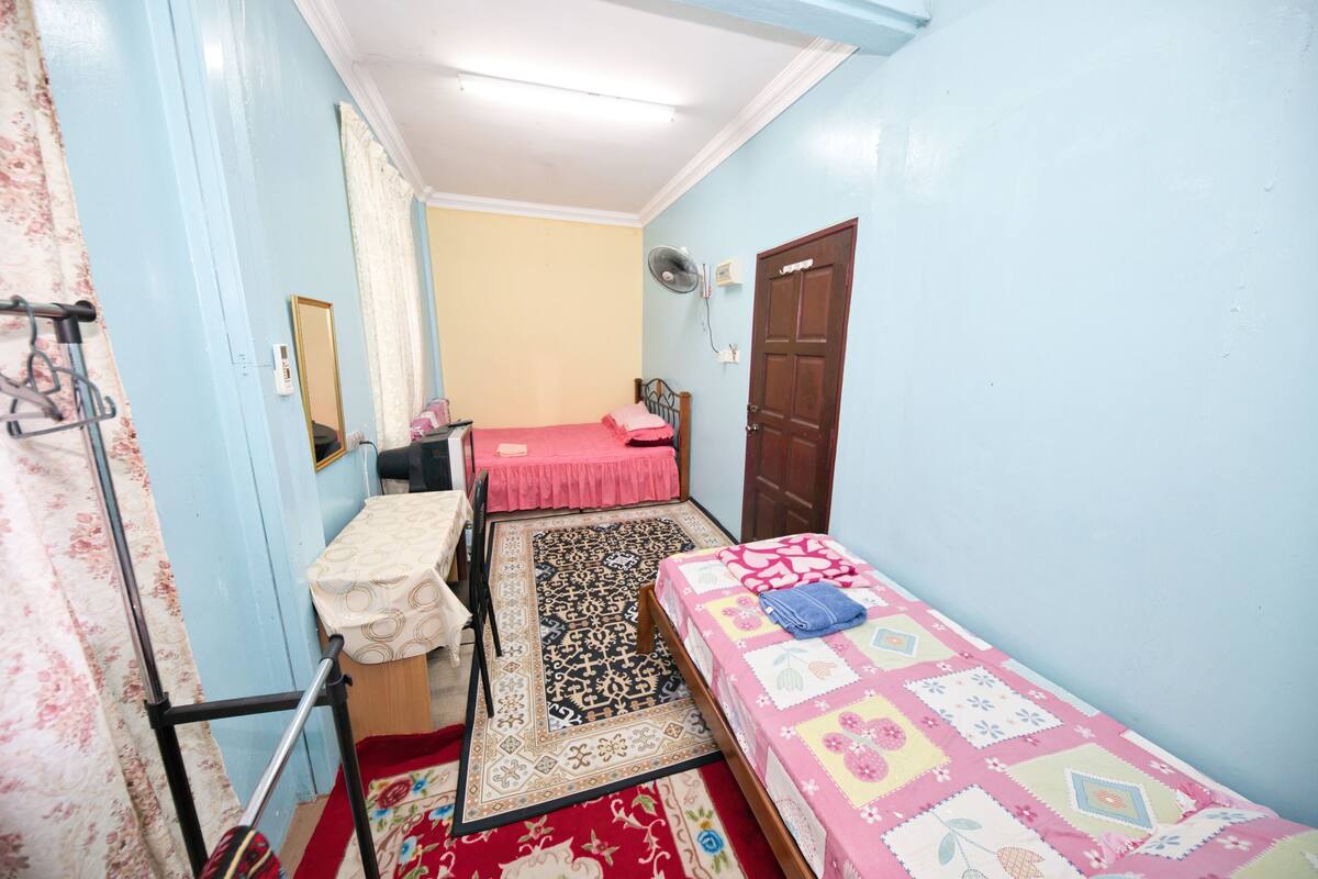 A small, well-lit bedroom with a single bed, a desk, and a chair. There is a mirror on the wall, a door, and a fan on the ceiling.