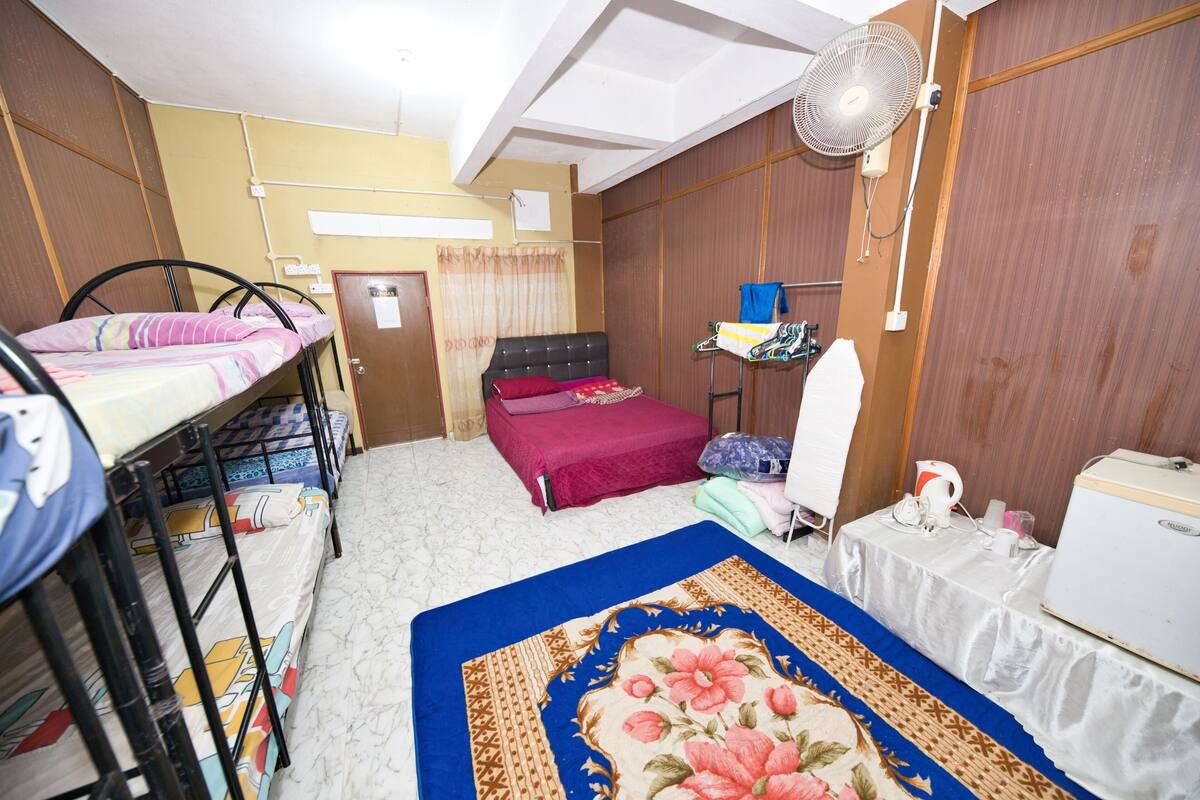 A room with bunk beds, a single bed, a fan, a table with a kettle, and a refrigerator.