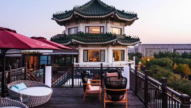 Un bâtiment traditionnel chinois, doté d'un toit végétalisé et de murs blancs, avec une terrasse en bois agrémentée de chaises et de tables en osier, et offrant une vue sur le paysage urbain en arrière-plan.