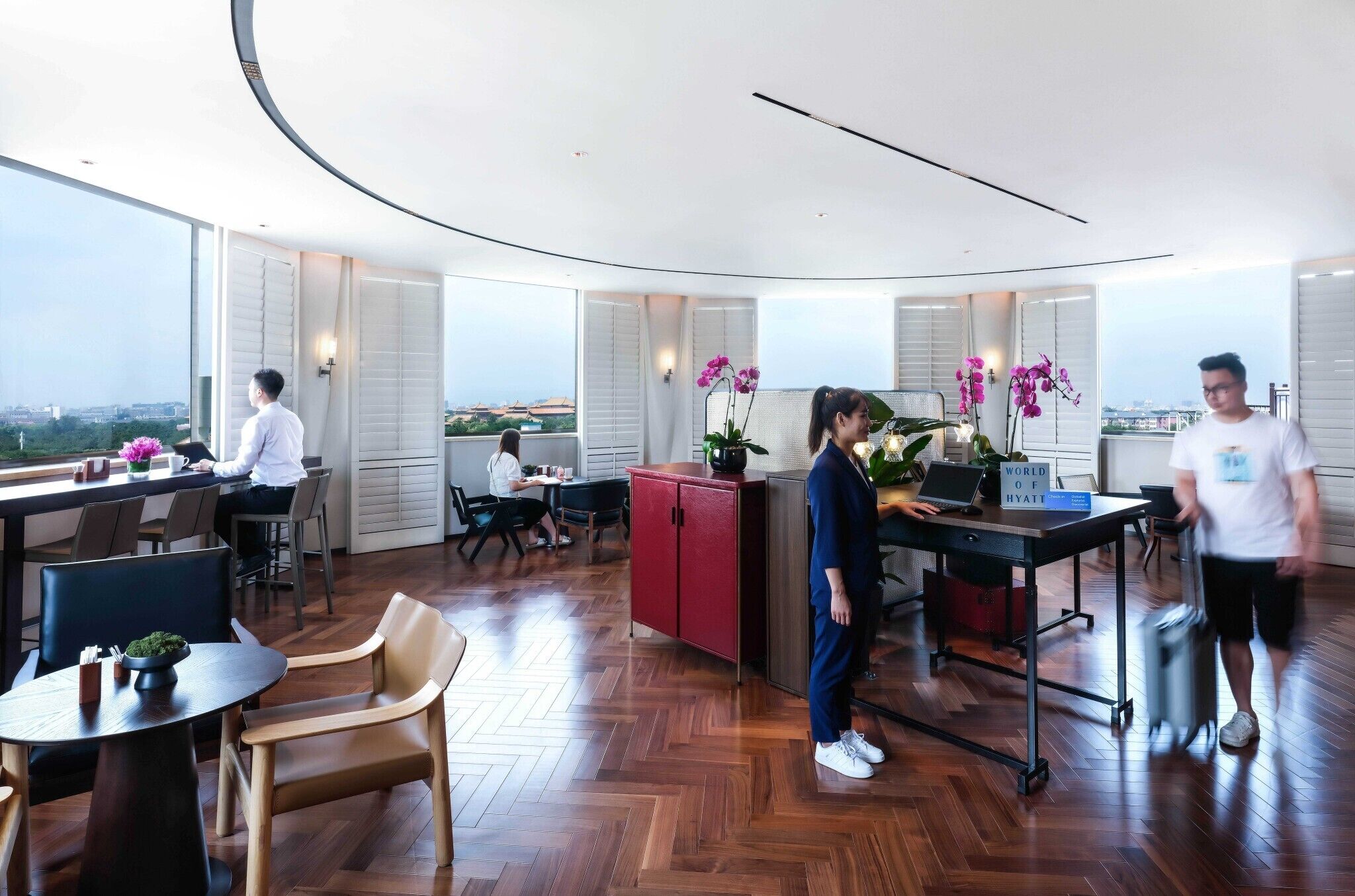 Un moderno vestíbulo de hotel con techo curvo, suelo de madera y ventanas amplias que ofrecen vistas de la ciudad. Hay varias mesas y sillas, y se observa un armario rojo.