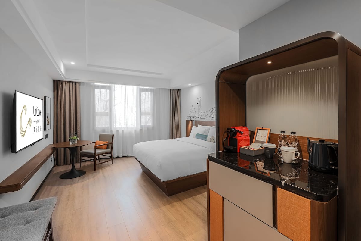 Una habitación de hotel con una cama, un escritorio con una silla, una pequeña mesa con una planta, una cafetera y un cuadro en la pared.