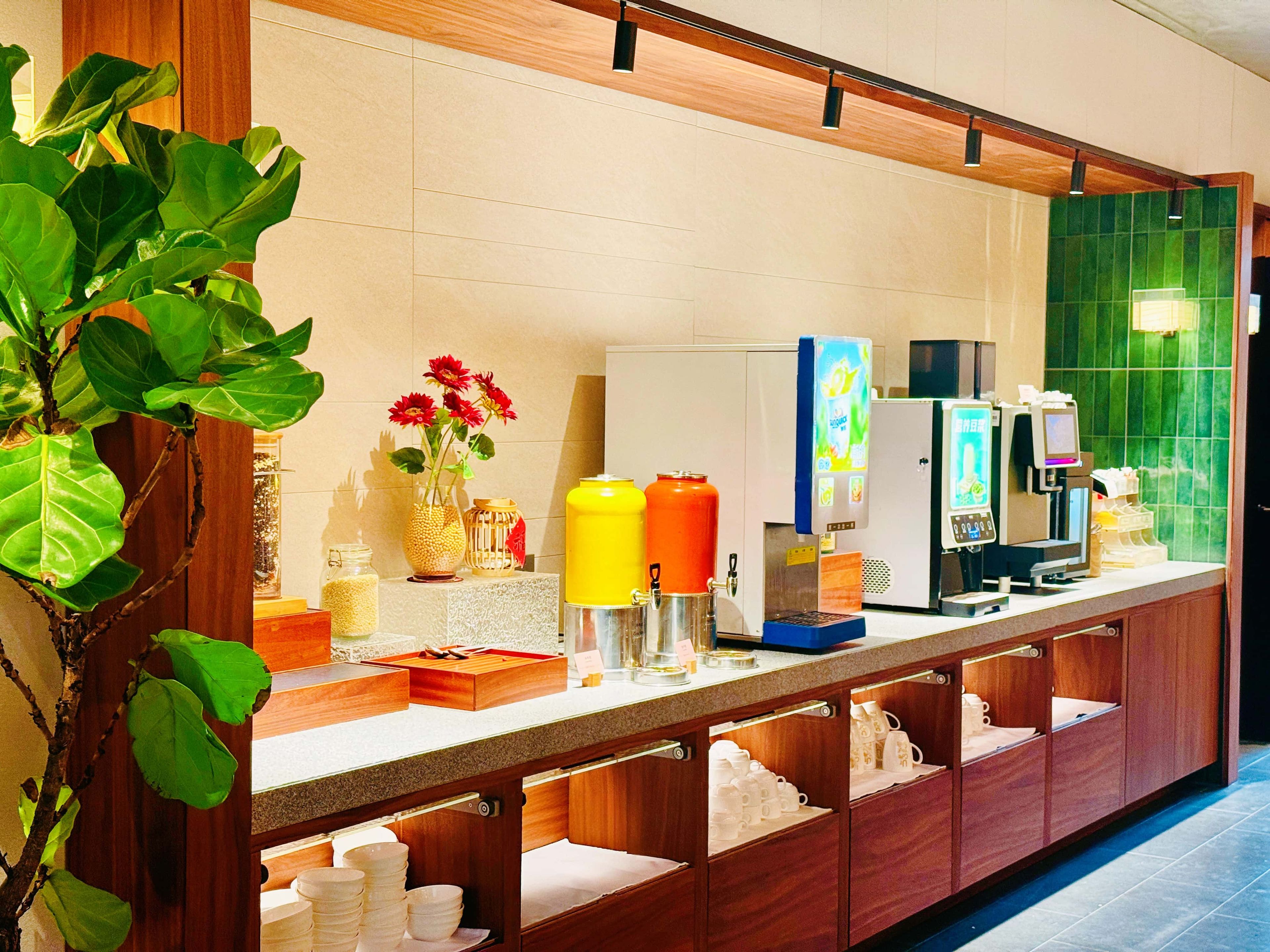 A hotel reception area with a counter, beverage dispensers, a refrigerator, and a green tiled wall.