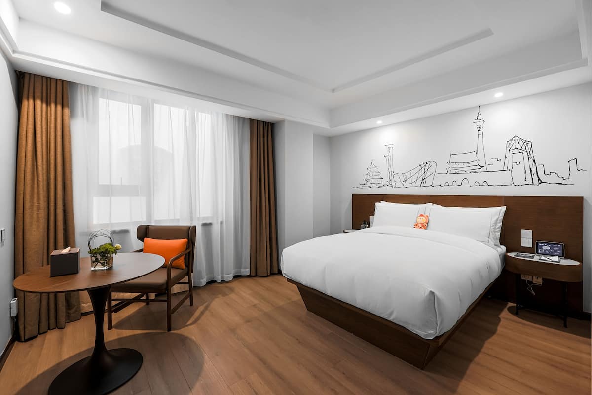 Habitación de hotel con cama, escritorio y silla, una mesita con un florero, y un mural urbano en la pared.