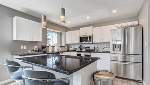 A modern kitchen with white cabinets, stainless steel appliances, and a marble island with bar stools. - Casa De Claudia (Colorado Springs)