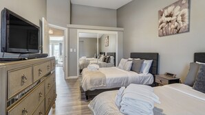 A hotel room with a large flat-screen TV, a dresser with drawers, two beds with neatly folded towels, a bedside table with a lamp, and a painting on the wall. - Casa De Claudia (Colorado Springs)