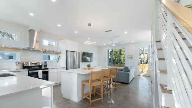 Una cocina moderna con armarios blancos, una isla central con taburetes altos y una zona de comedor con sofá.