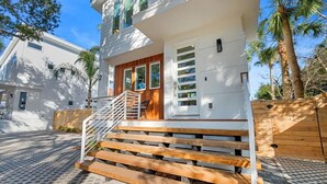 Una casa moderna de dos pisos con una escalera de madera que lleva a una puerta principal, rodeada de un área pavimentada y palmeras.