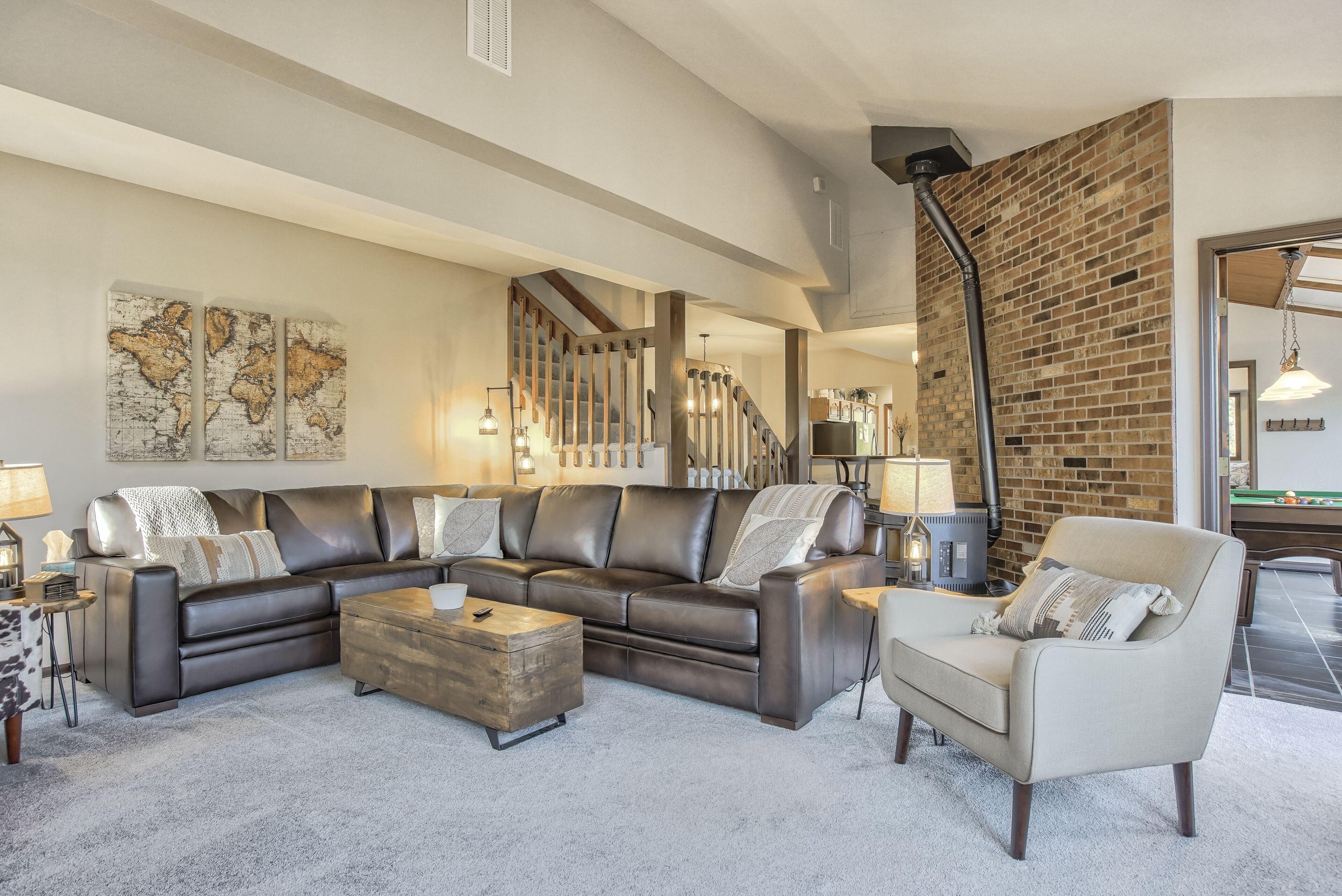 A spacious living room with a large sectional sofa, a wooden coffee table, and a beige armchair. A brick accent wall and a staircase are visible in the background.