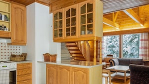 A wooden kitchen with a central island, a dining area with a sofa and chairs, and a view of snow-covered trees through large windows.