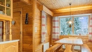 A wooden cabin interior with a dining table and benches, a window with a view of snow-covered trees, and a hanging light fixture.